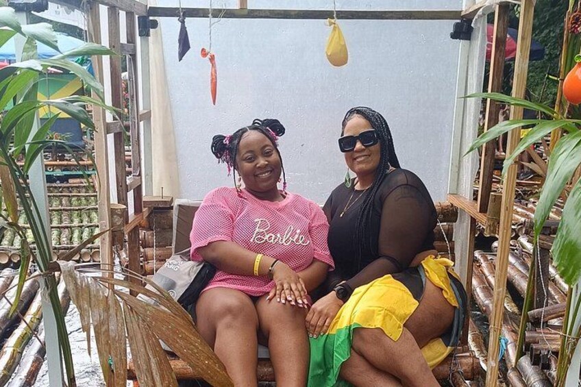 Two people sitting on a wooden swing, surrounded by plants and a wooden structure. 4