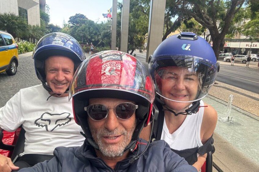 Drie personen op een motor, met helmen, met een stadsstraat en gebouwen op de achtergrond. 4