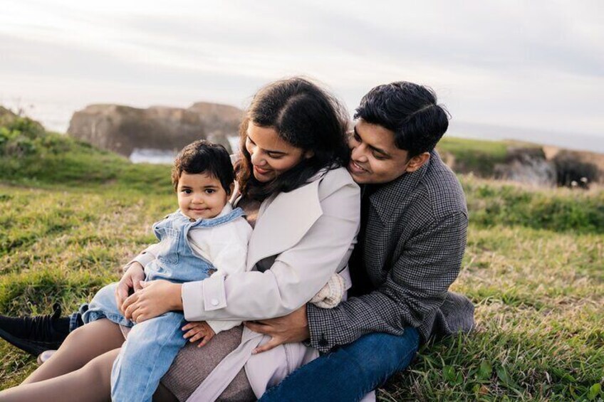 A family of three sitting on grass with a rocky coastline in the background. 2