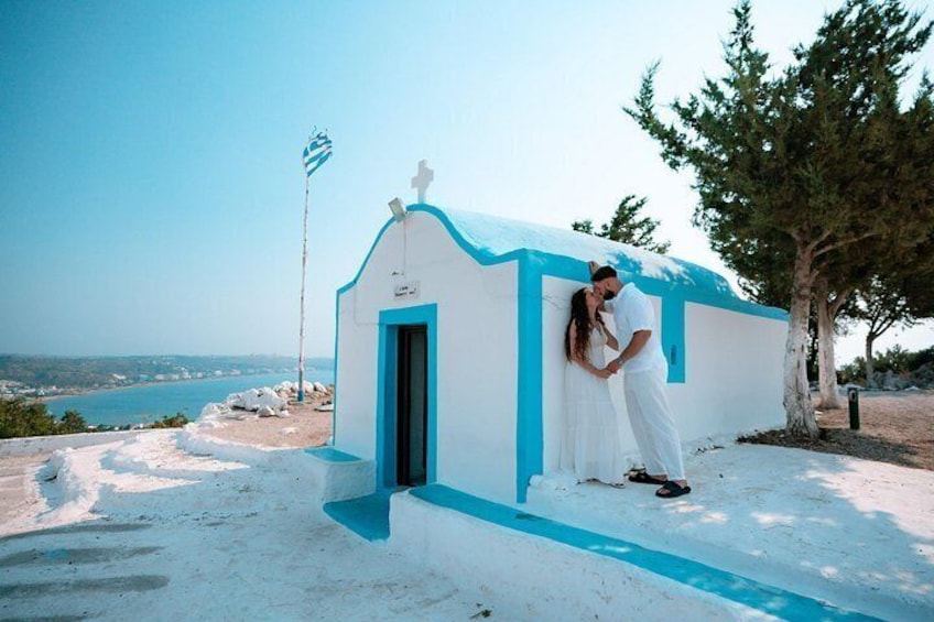 A couple embracing near a white and blue building with a cross on top, overlooking a coastal landscape. 5