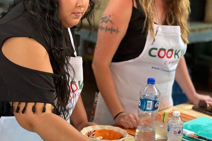 Due persone con grembiuli con la scritta 'COOK', sedute a un tavolo con una bottiglia d'acqua e un piatto di cibo. 5