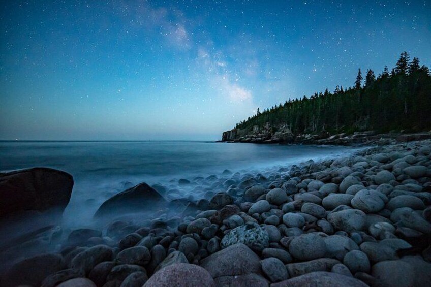 A starry night sky over a rocky beach with a forest in the background. 2