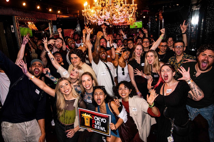 A crowded indoor event with a chandelier and a sign reading 'TOKYO PUB CRAWL'. 1