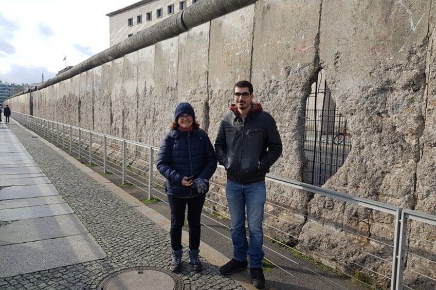 Two people standing by a weathered stone wall with a metal fence, a cobblestone path, and a building in the background. 3