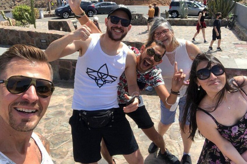 A group of people posing for a photo in an outdoor setting with a stone wall and parked cars in the background. 3