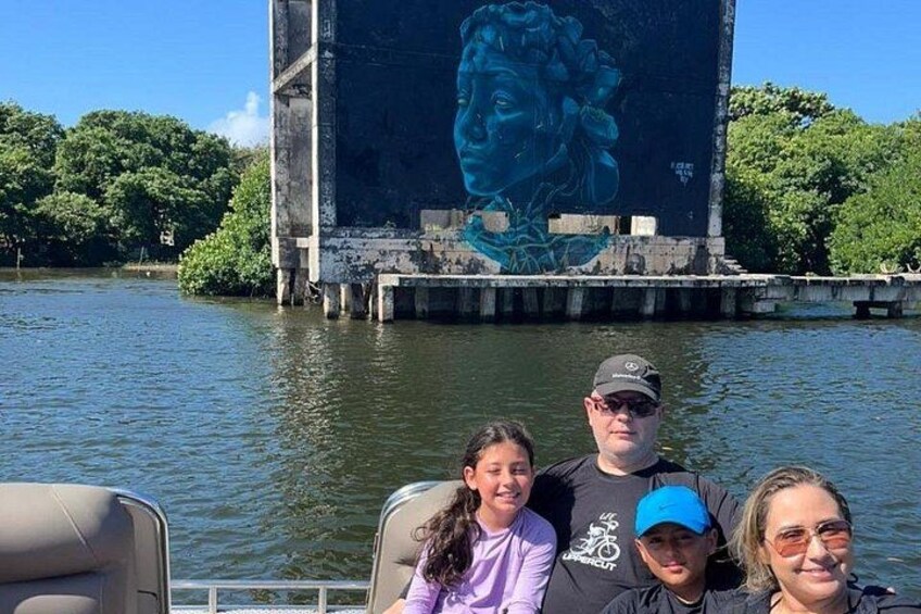 A group of people on a boat near a large mural of a woman's face on a building by the water. 4