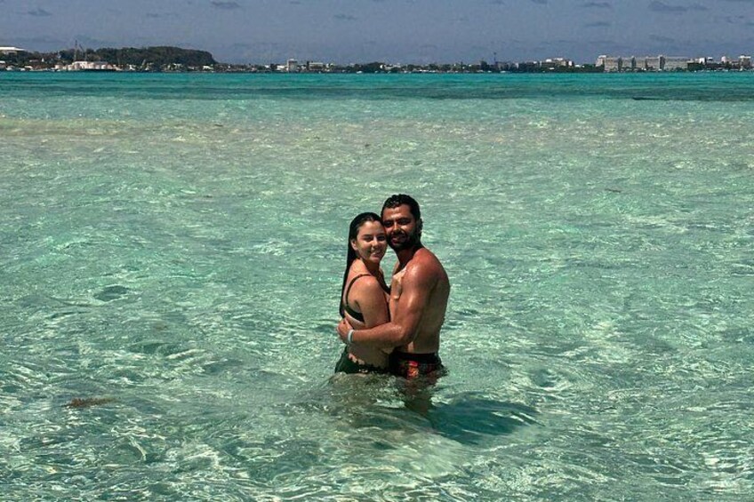 A couple embracing in shallow clear water with a distant shoreline and buildings. 3