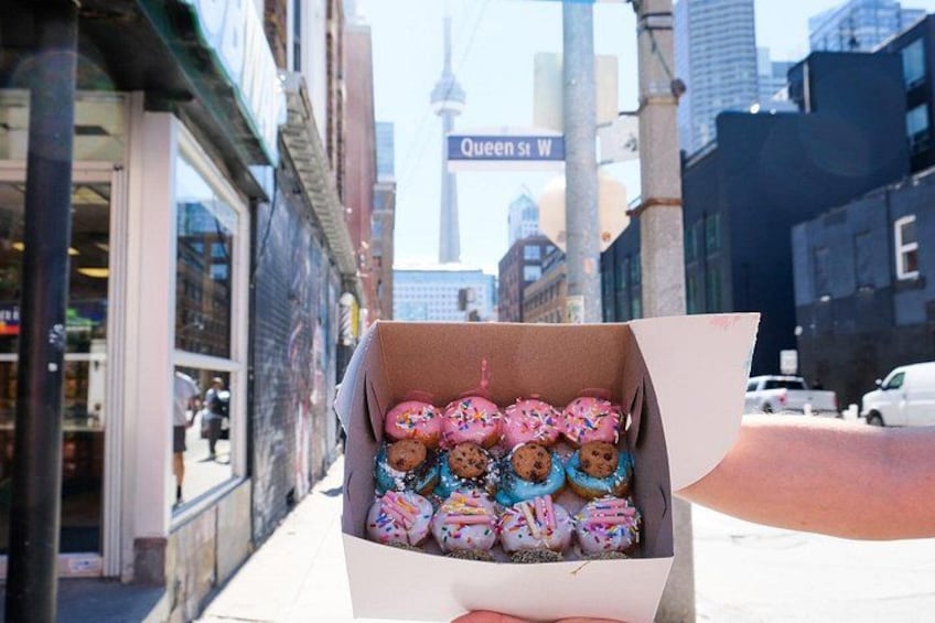 A box of colorful donuts on a sidewalk with a street sign and buildings in the background. 3