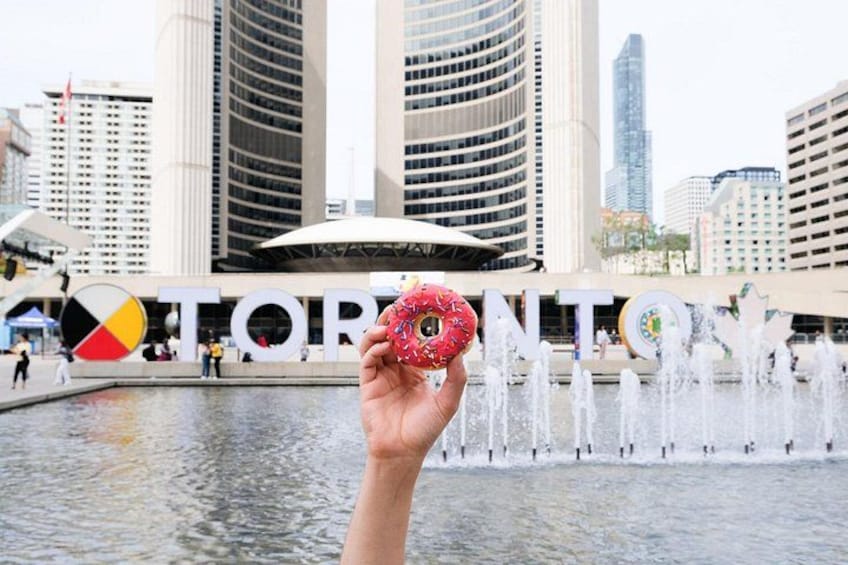 En hånd holder en doughnut foran en springvand, med ordet 'TORONTO' i baggrunden. 2
