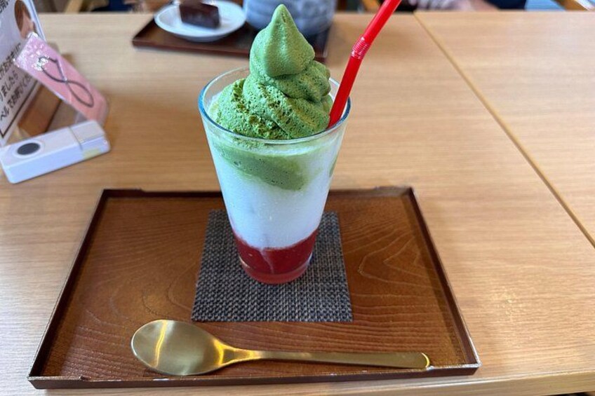 Un dessert composé de plusieurs couches, surmonté d’une garniture verte, avec une paille rouge et une cuillère dorée, le tout présenté sur un plateau en bois. 2