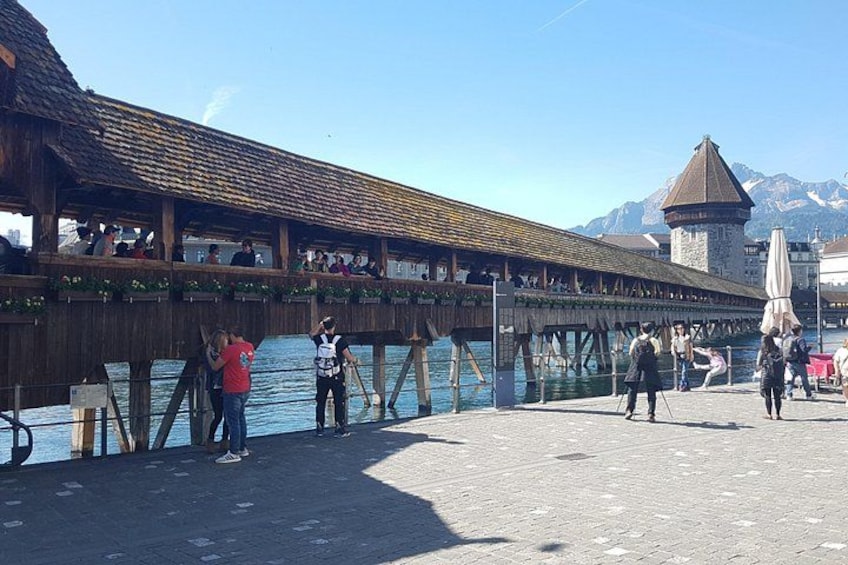 Een houten steiger die over het water loopt, met mensen die lopen en zitten, een berg op de achtergrond en een torenachtige constructie. 3