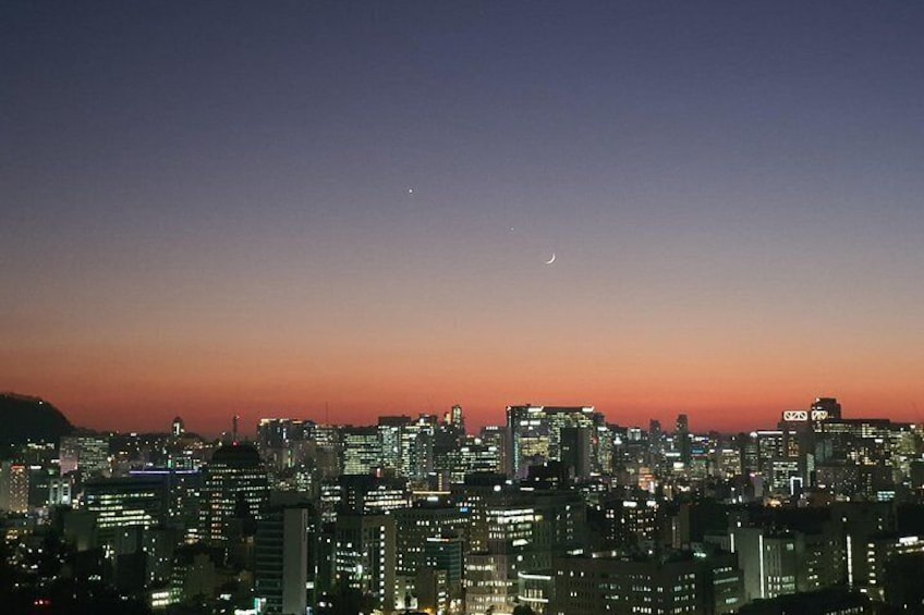 Une vue sur les gratte-ciel d’une ville au crépuscule, avec des bâtiments illuminés et un croissant de lune dans le ciel. 3