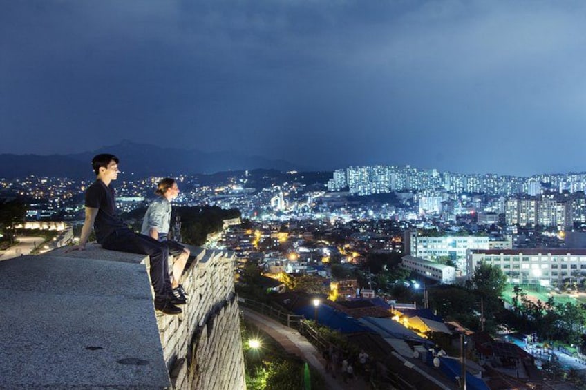 Twee mensen zitten op een randje en kijken uit over een stadsgezicht in de nacht. 1