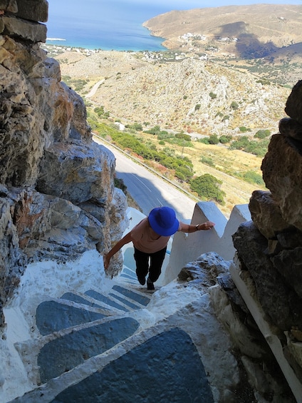 Una persona baja por una estrecha escalera de piedra, con un sombrero azul, con vistas a un paisaje costero con edificios y vegetación. 2