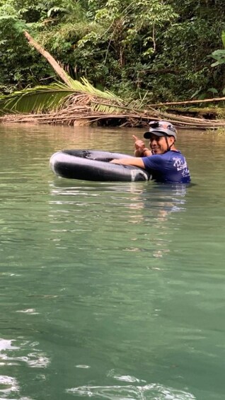 En person i en blå skjorta och hjälm flyter på en flotte i en flod, omgiven av tät vegetation. 2