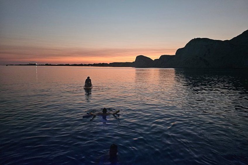 A sunset over a calm body of water with people swimming and a large rock formation in the distance. 4