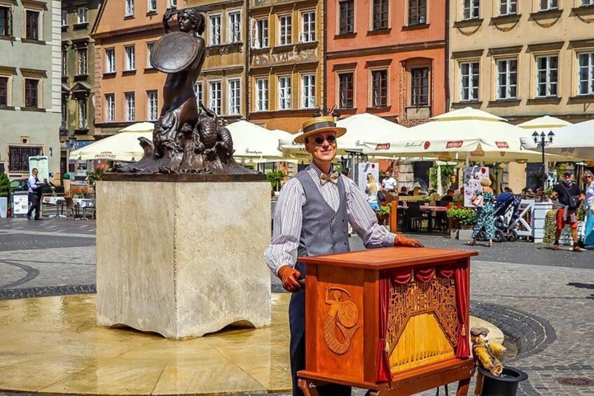 A person in a striped shirt and hat standing next to a wooden box with a red curtain, in front of a statue and buildings. 2