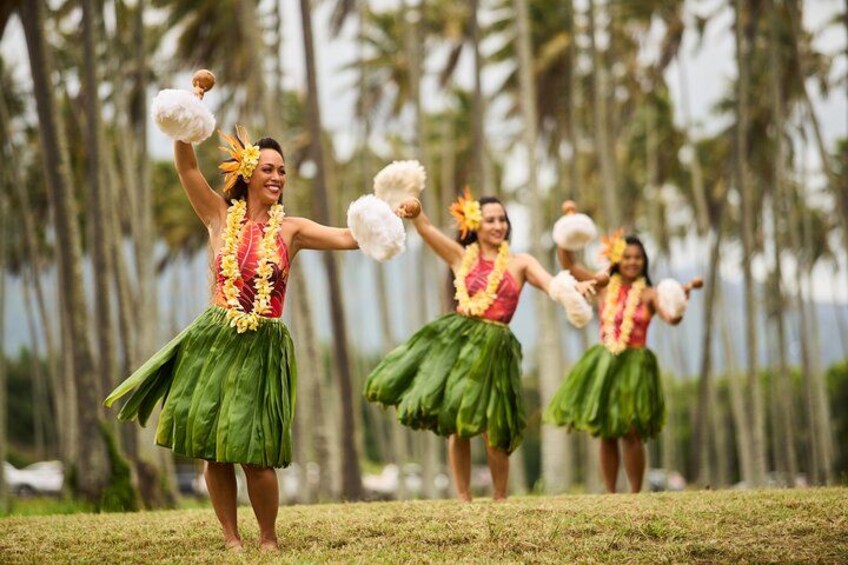 Un grupo de personas bailando una danza tradicional con faldas verdes y blusas blancas, sosteniendo abanicos blancos, en un área con césped y palmeras al fondo. 1