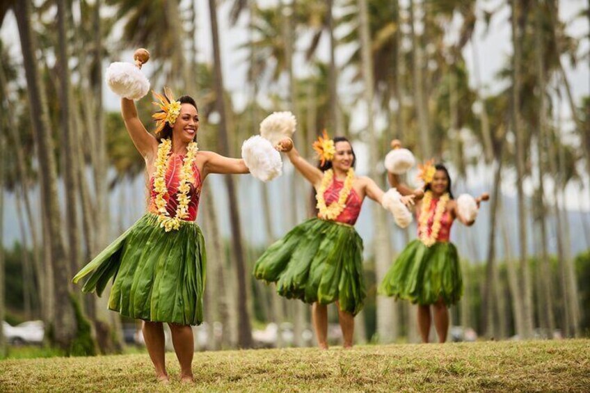 Three people in green skirts and leis, holding pom-poms, performing in front of palm trees. 1