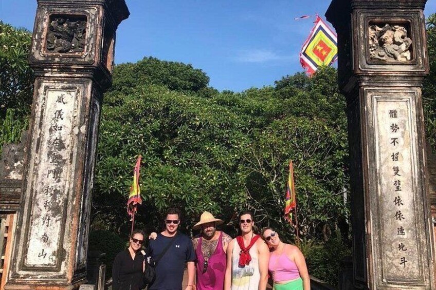 A group of people standing in front of an ornate gate with inscriptions, surrounded by lush greenery and flags. 4