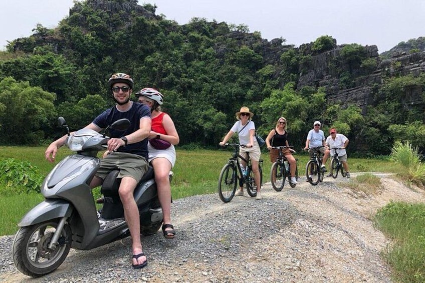 Un grupo de personas andando en patinete y bicicletas por un camino de grava, con zonas verdes y colinas rocosas al fondo. 2