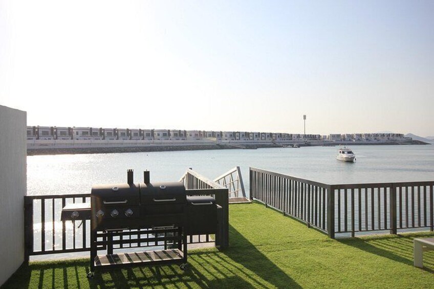 A balcony with a grill, a bench, and a view of a waterfront with a boat and a distant shoreline. 3
