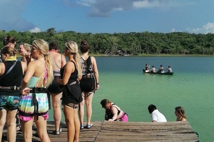 A group of people on a wooden dock by a calm lake, with a small boat in the distance. 2