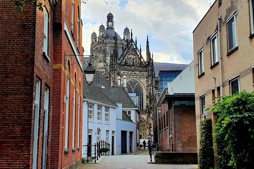 A narrow street with historic buildings, a church with a prominent spire, and a modern building in the background. 1