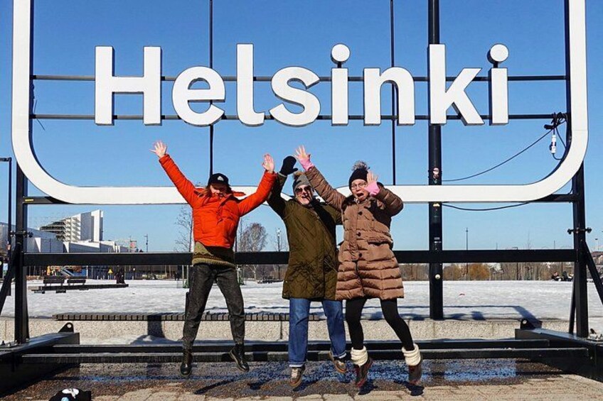 Drie personen poseren voor een groot bord waarop 'Helsinki' staat. 3