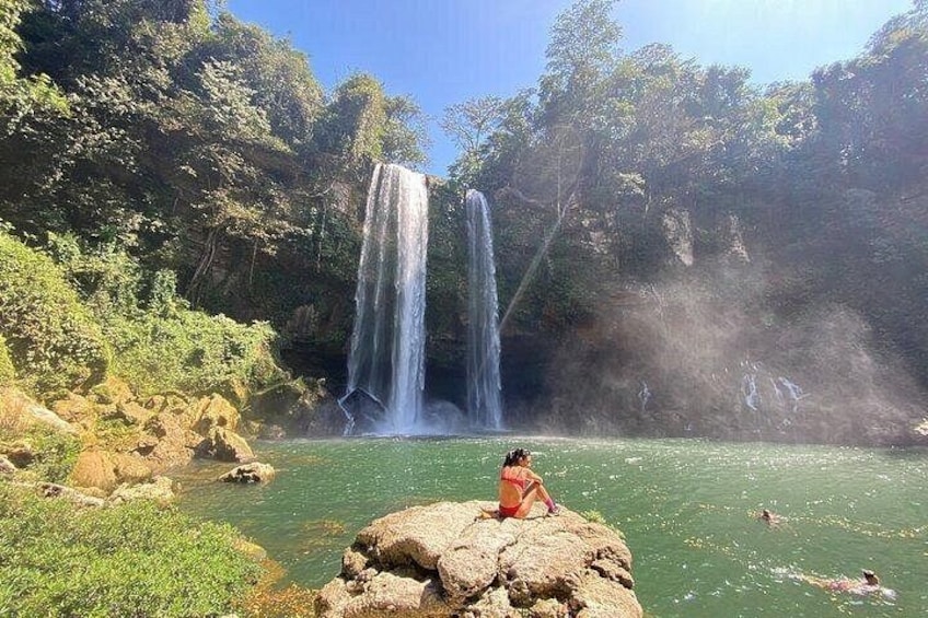 A waterfall, a person sitting on a rock, and people swimming in a body of water. 5