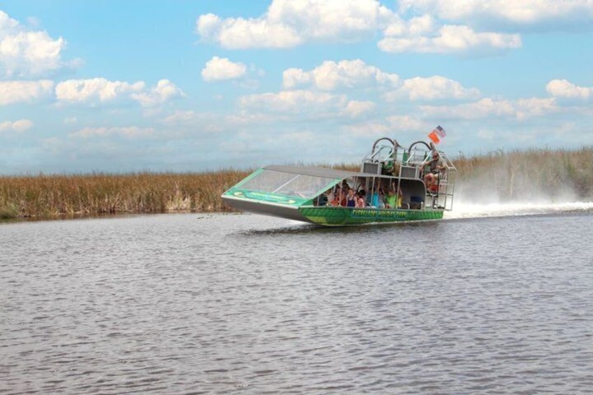 Un'imbarcazione verde con passeggeri che naviga sull'acqua, con una riva erbosa e cielo sereno. 1