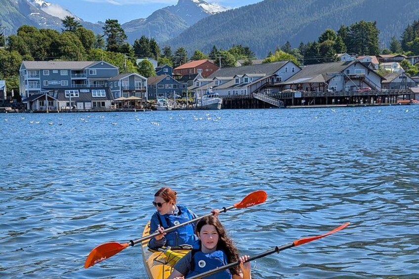 Two people kayaking on a calm lake with houses and mountains in the background. 3