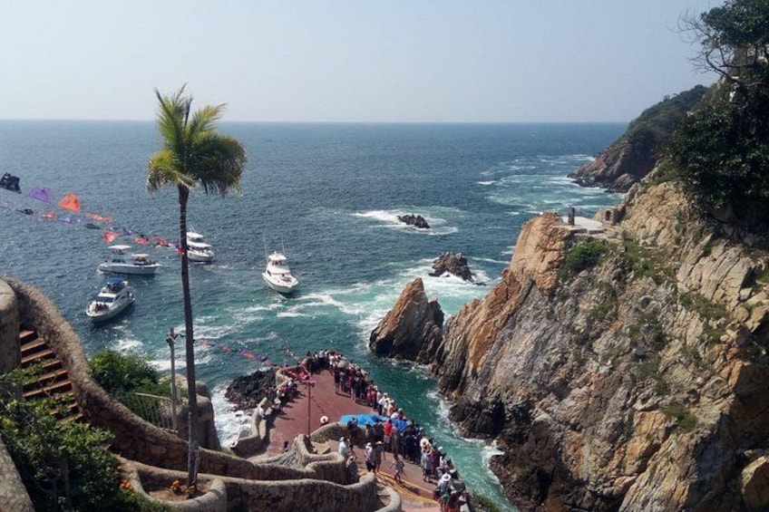 A coastal scene with boats, a rocky cliff, and a group of people on a path. 1