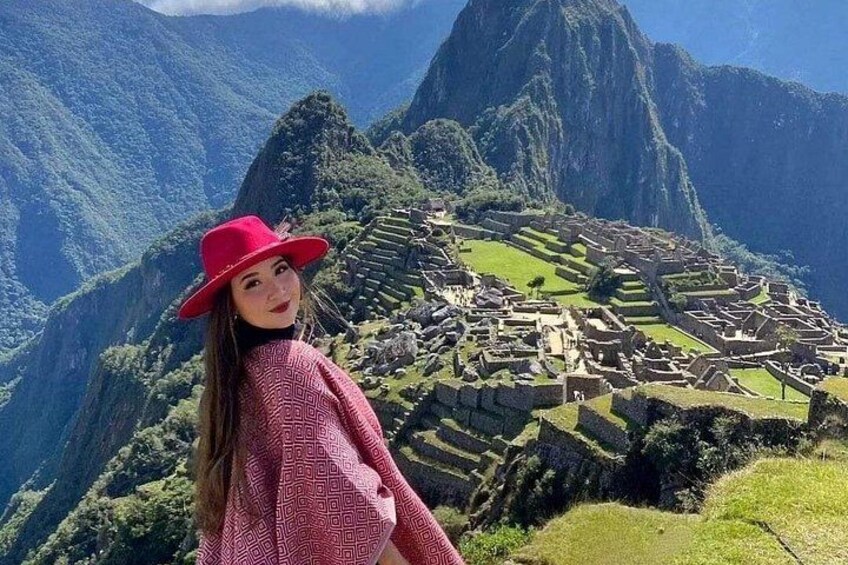 Une personne portant un chapeau rouge et un haut à motifs se tient devant le Machu Picchu, avec ses terrasses agricoles et ses ruines antiques. 1