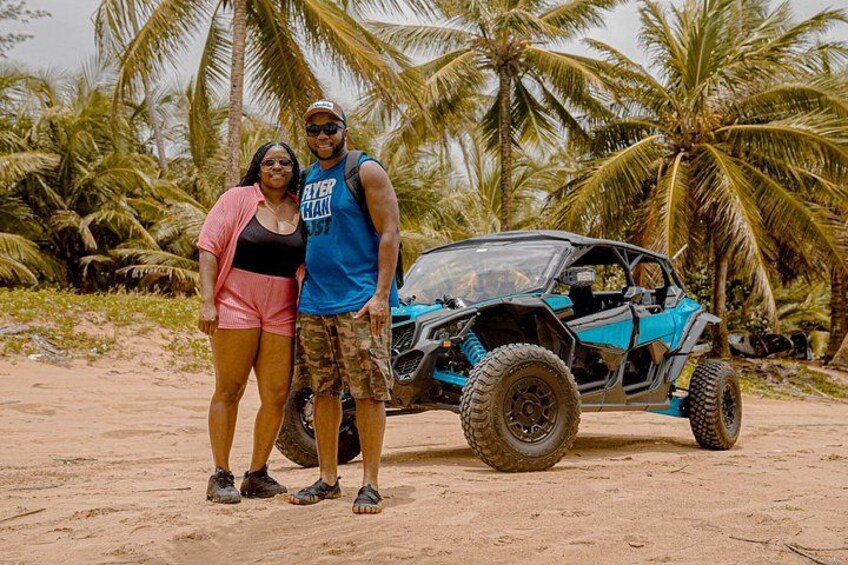 Deux personnes se tiennent devant un véhicule tout-terrain bleu sur une plage de sable, avec des palmiers en arrière-plan. 3
