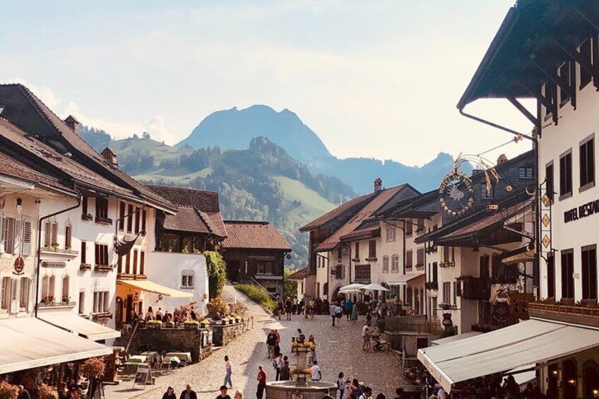 En kullerstensgata i en europeisk by med traditionella byggnader, ett klocktorn och berg i bakgrunden. 4