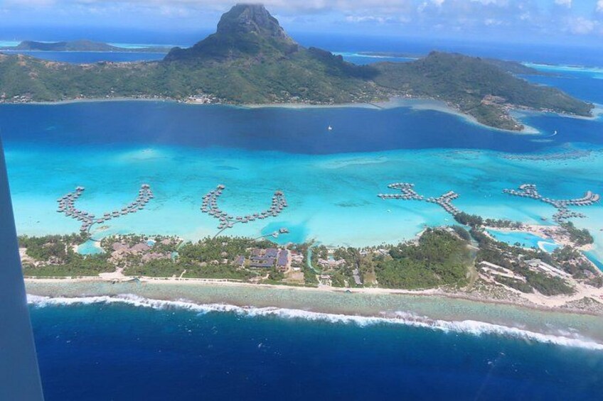 An aerial view of a tropical island with clear turquoise waters, a prominent mountain peak, and a series of circular structures on the beach. 4