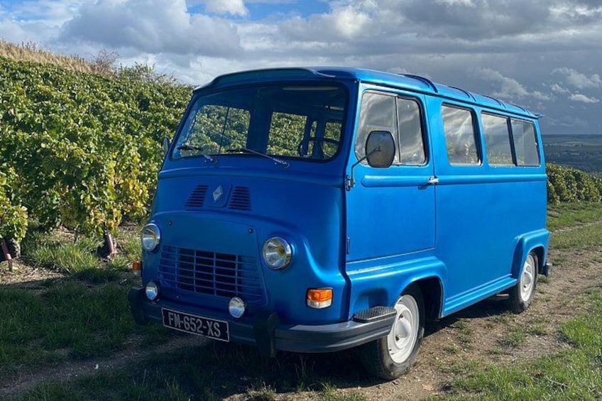 A blue vintage van parked on a dirt path with vineyards in the background. 2