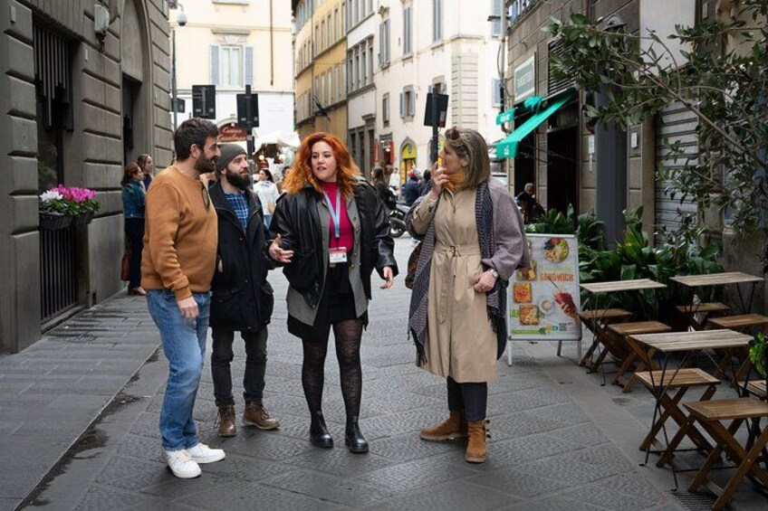 Un grupo de personas caminando por una calle urbana con edificios y zonas de estar al aire libre. 1