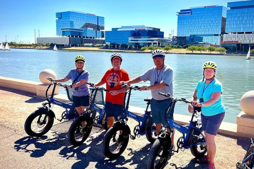 Fyra personer på cyklar vid en strandpromenad, med moderna byggnader i bakgrunden. 2