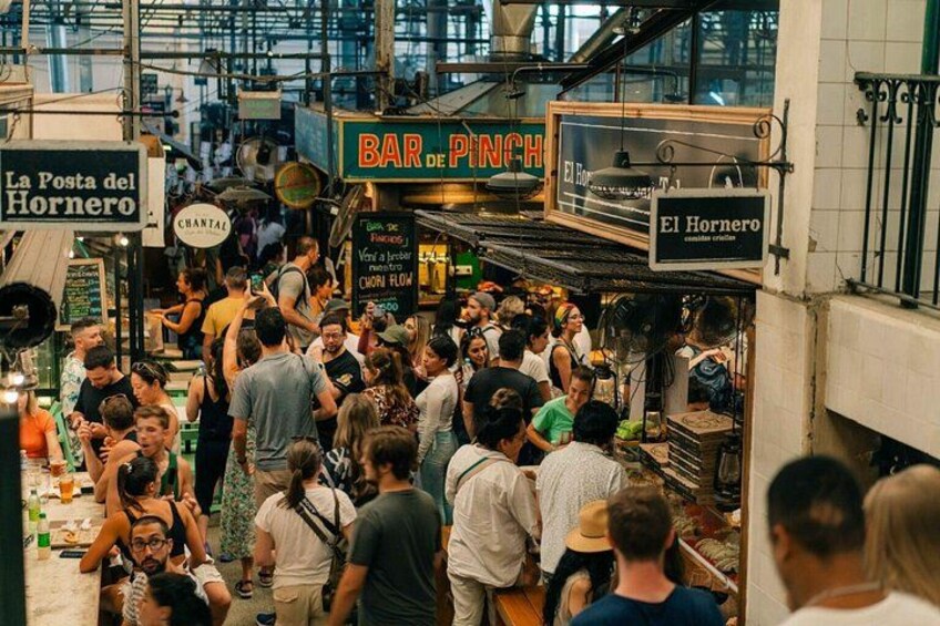 Un mercado cubierto concurrido, con letreros que indican 'La Posta del Hornero' y 'BAR DE PINCHE'. 3