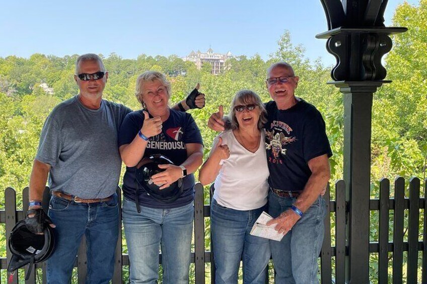 Four people standing on a wooden deck with a metal lamp post, trees, and a building in the background. 4