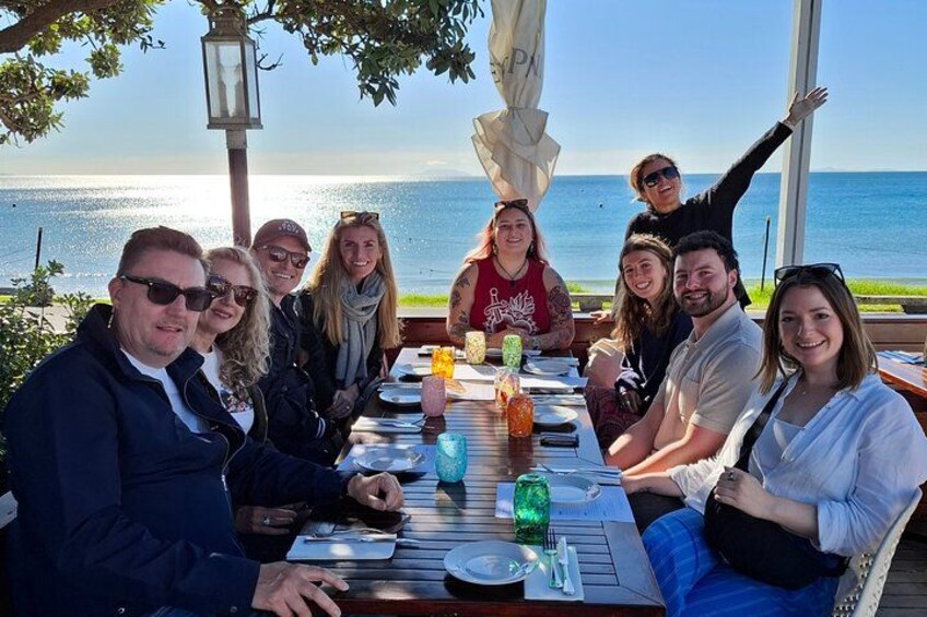 En gruppe mennesker spiser ved et bord udendørs med udsigt over havet. 1