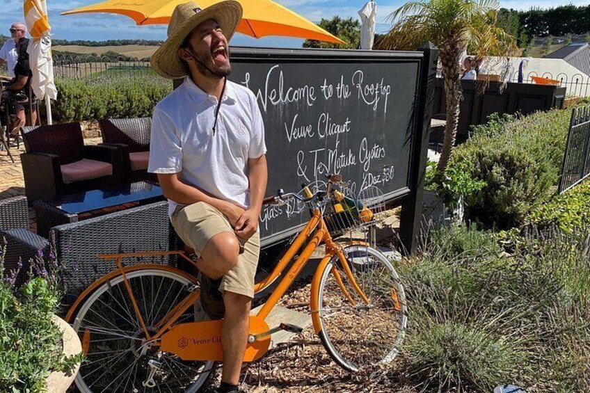 A man sitting on a vintage orange bicycle with a sign in the background that reads 'Welcome to the Rooftop Vineyard.' 2