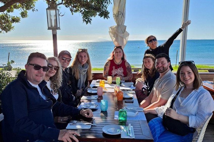 Un grupo de personas cenando en una mesa al aire libre con vista al océano. 1