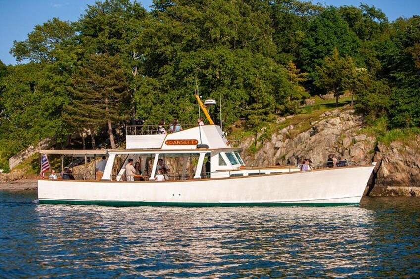 A white boat named 'Gansett' on calm water with a rocky shore and dense trees in the background. 2