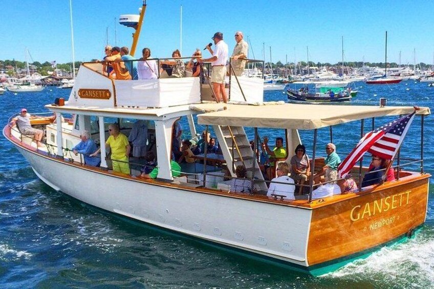 A boat named Gansett sailing on water with people on board and a marina in the background. 1