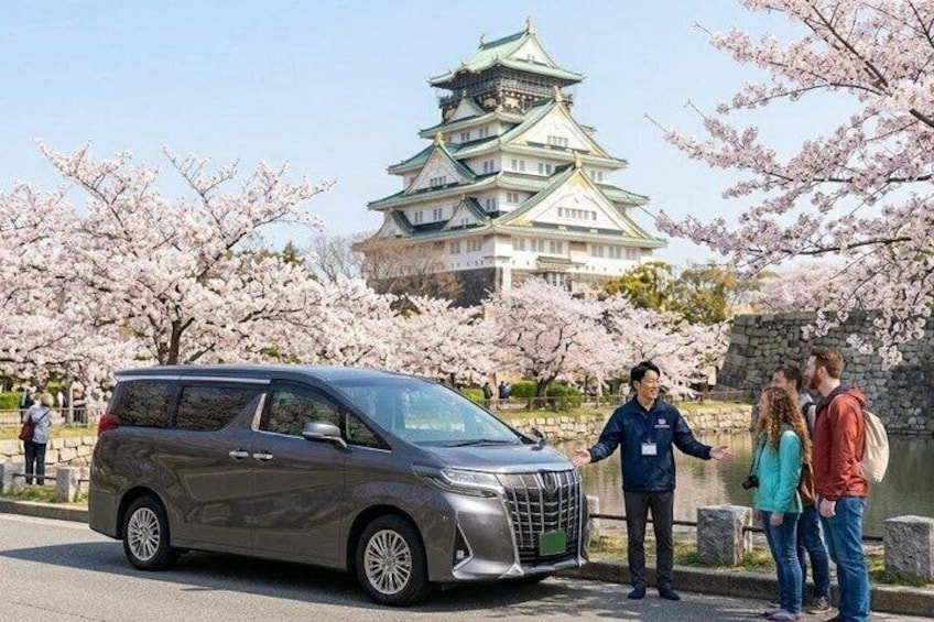A silver minivan parked in front of a traditional Japanese castle with cherry blossoms. 1