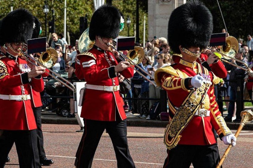 Een groep bewakers in rode uniformen met zwarte hoeden die messing blasinstrumenten bespelen. 2