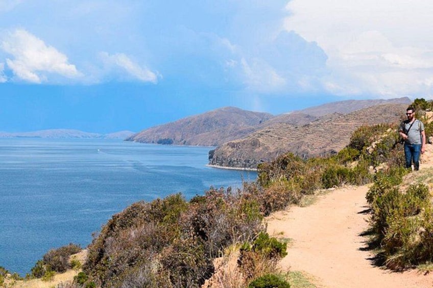 Un sendero costero con una persona caminando, rodeado de colinas y una masa de agua. 3